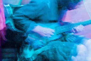 A blue and violet live performance photograph showing Anuar playing guitar